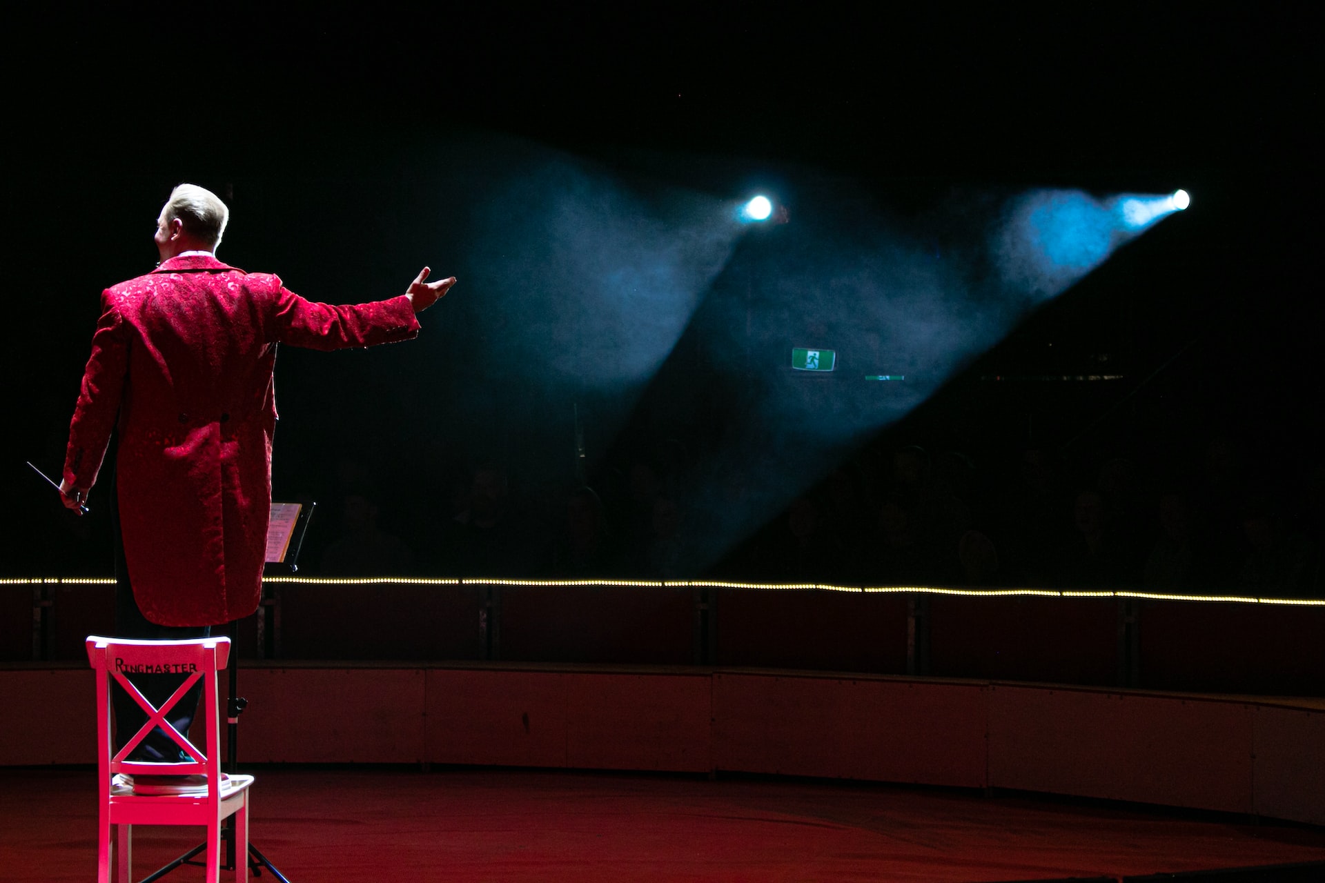 a ringmaster in a red suit standing on a chair in front of spotlights