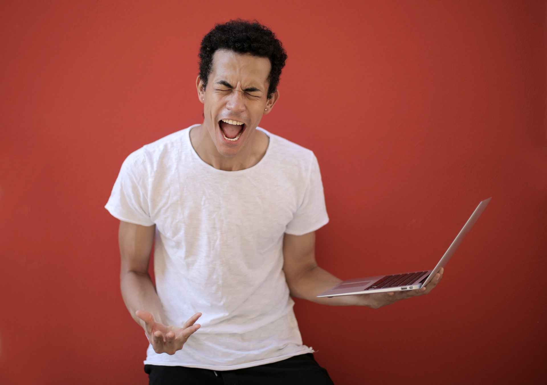 person in white t shirt holding laptop screaming in front of red background