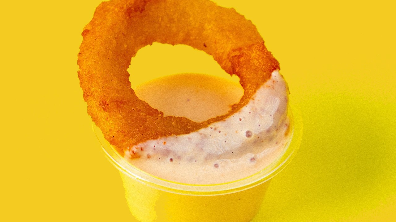 an onion ring being dipped into a plastic container of sauce, on yellow background